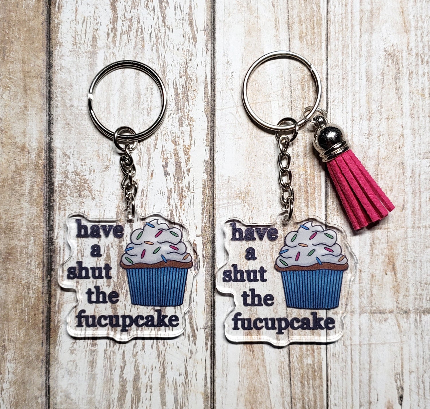 A photo of two acrylic keychains. They have cupcakes on them. Text next to cupcakes reads 'have a shut the fucupcake'