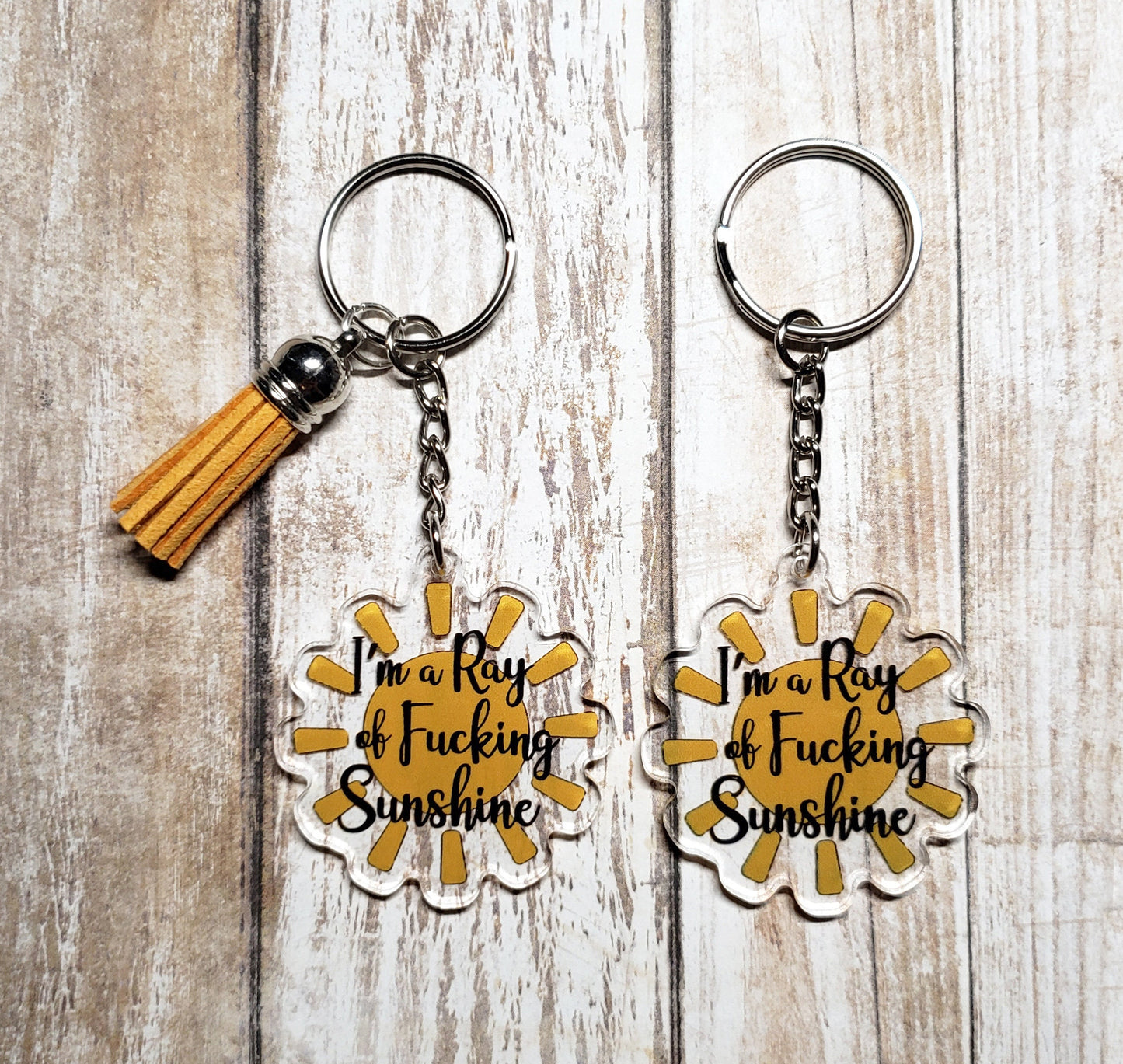 A photo of an acrylic keychain. The keychain is a yellow sunshine. Text on keychain reads 'I'm a Ray of Fucking Sunshine.'