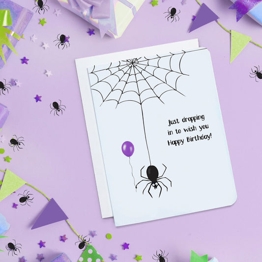 A photo of a white funny birthday card. It has a spider web at the top, a spider is hanging from it. It's holding a purple balloon. Text on card reads, 'Just dropping in to wish you Happy Birthday!'
