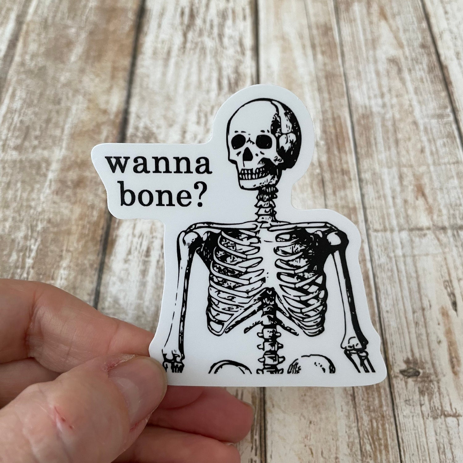 A photo of a vinyl sticker of cartoon skeleton. Text on sticker reads 'Wanna Bone?.'