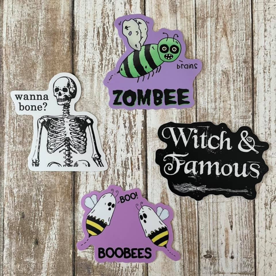 A photo of four vinyl halloween stickers. One a Skelton and text reads 'Wanna Bone?' Two a Zoo-bee. Three text sticker reads 'Witch & Famous.' Four, two bees with sheets on head. Text reads 'BooBees.'